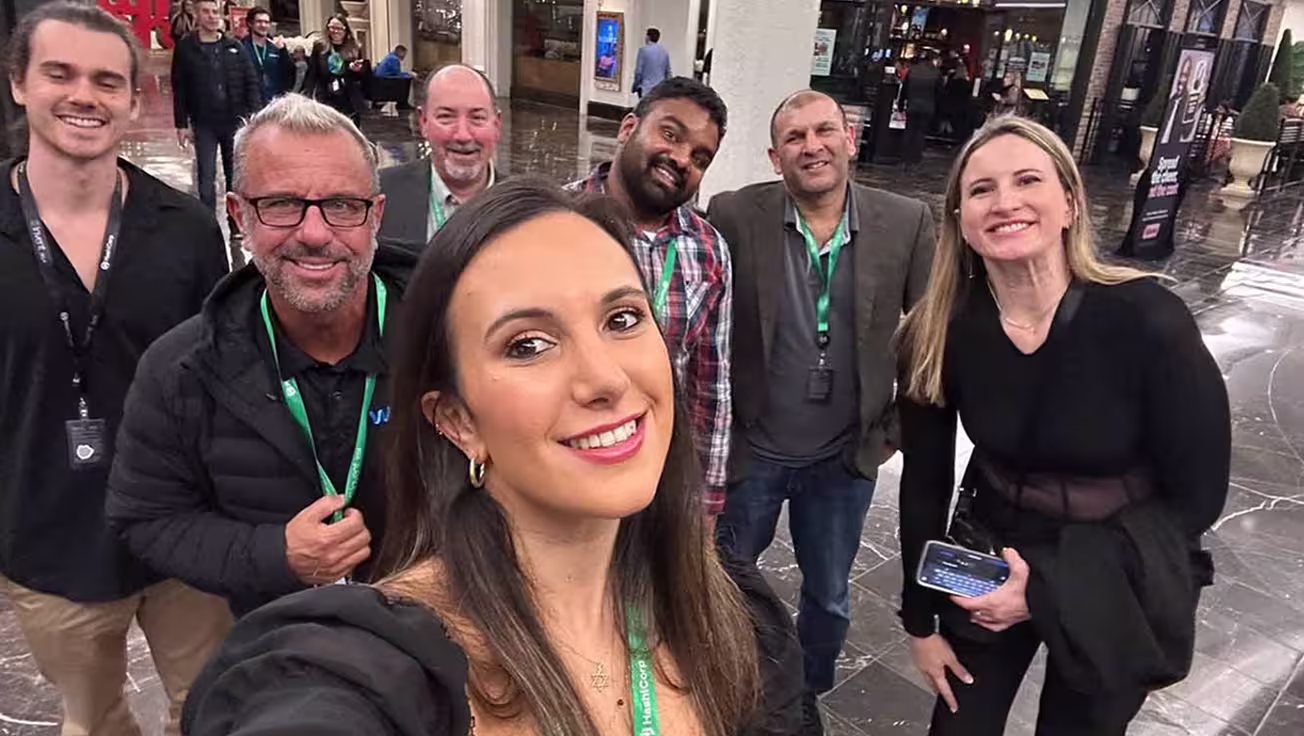 Group of seven smiling adults posing for a selfie indoors at a public event or conference.